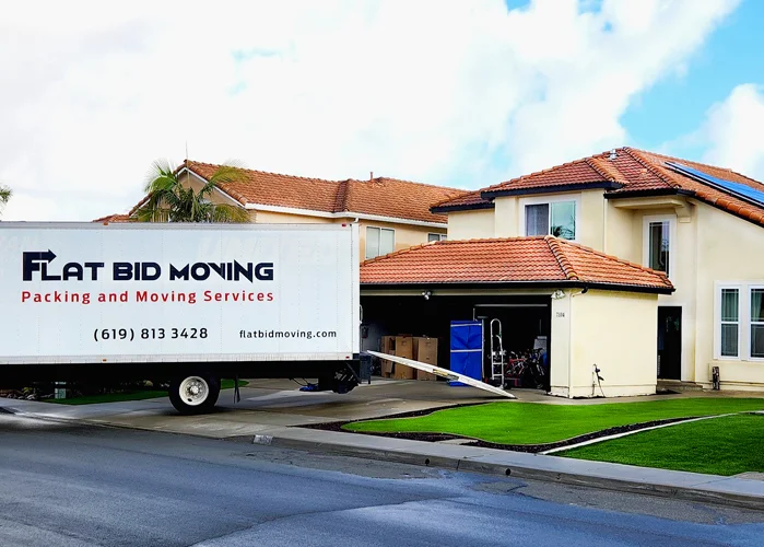 Flat Bid Moving LLC truck parked in front of a two-story home while movers load items from the garage.