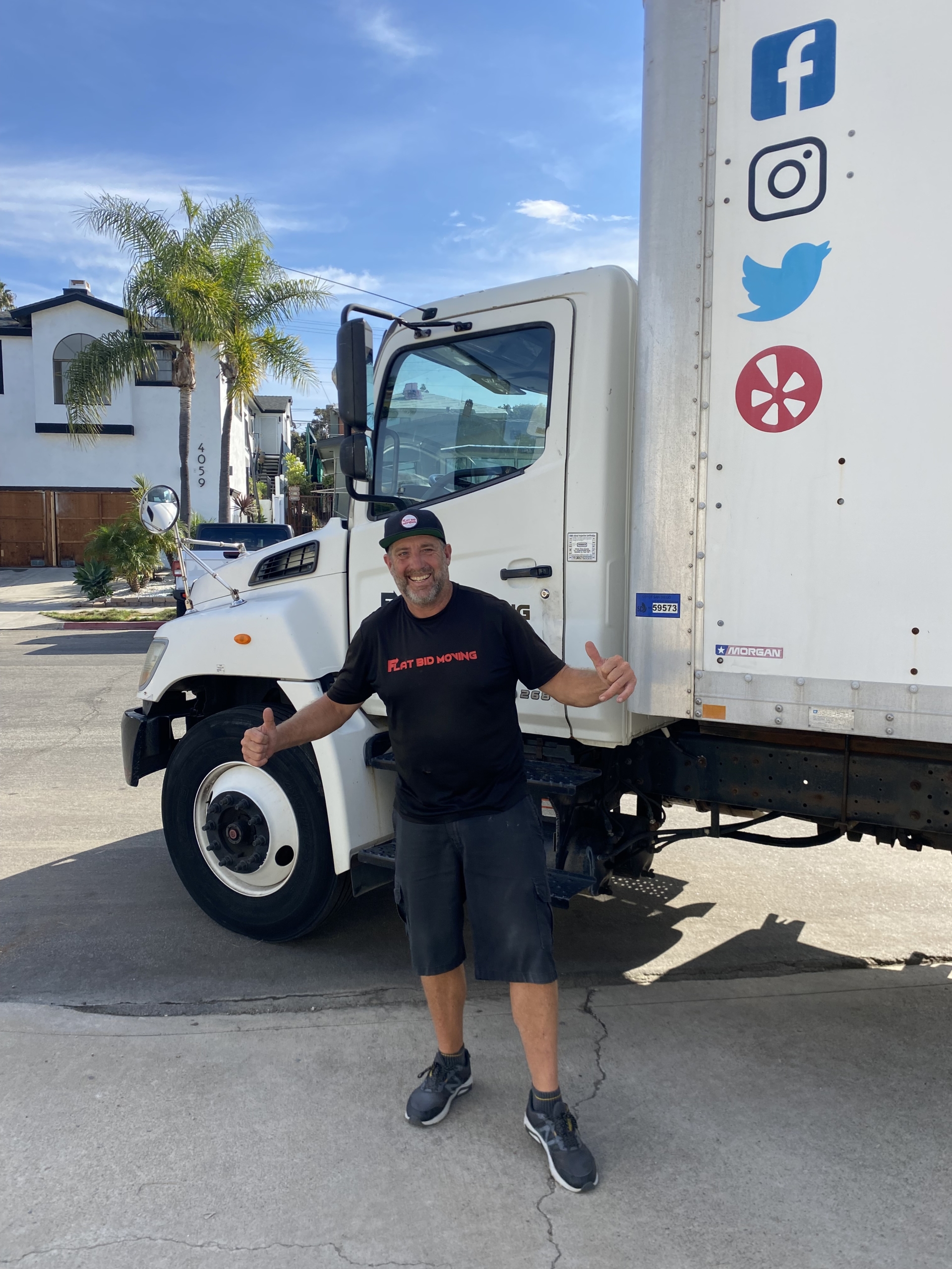 Flat Bid Moving LLC team member standing in front of a branded moving truck, giving a thumbs-up gesture in a residential neighborhood.