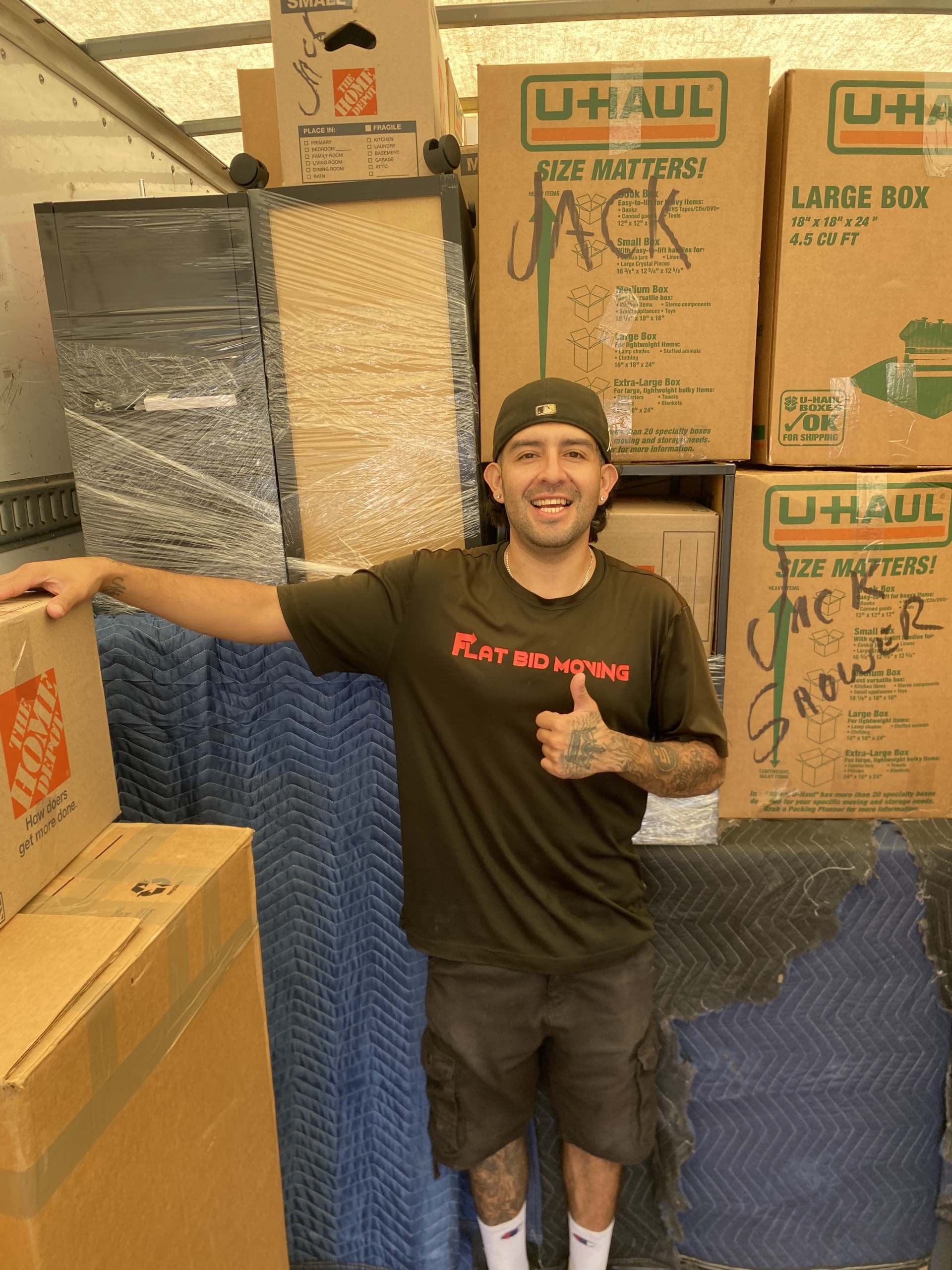 Flat Bid Moving LLC mover giving a thumbs-up while standing inside a packed moving truck filled with labeled boxes.