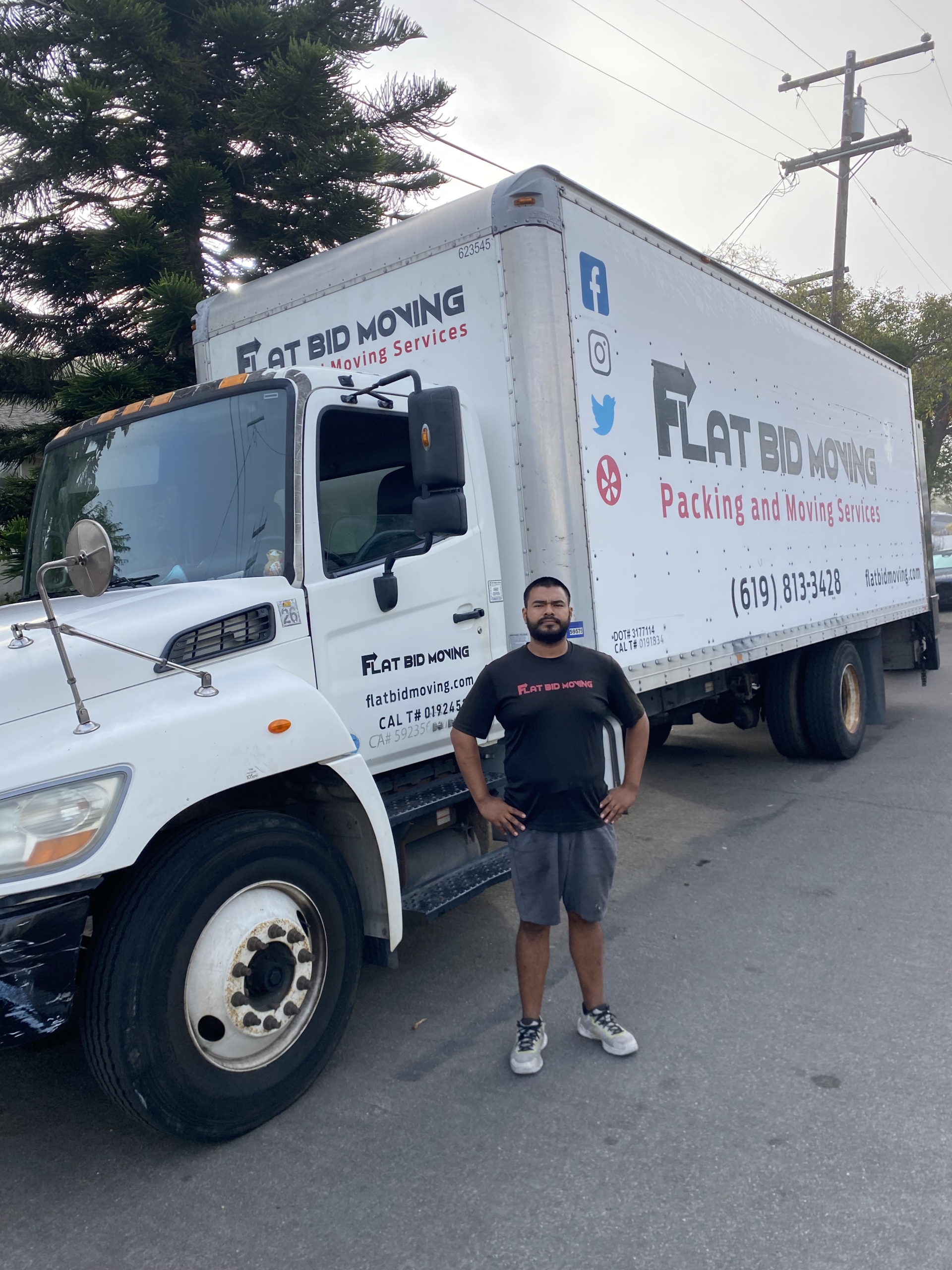 Flat Bid Moving LLC mover standing beside a large moving truck parked on a residential street.